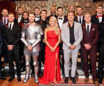 Group photo of "The Bachelorette Australia 2018" contestants in formal wear, with one in a knight costume, posing indoors.