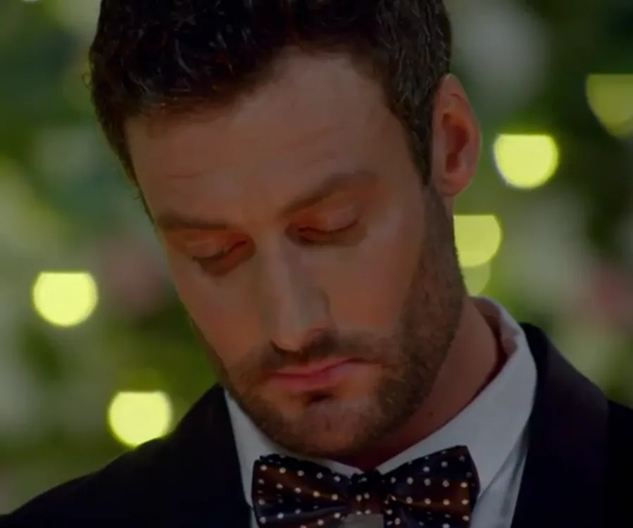 A man in a suit with a polka dot bow tie, looking down, set against a bokeh-lit background.
