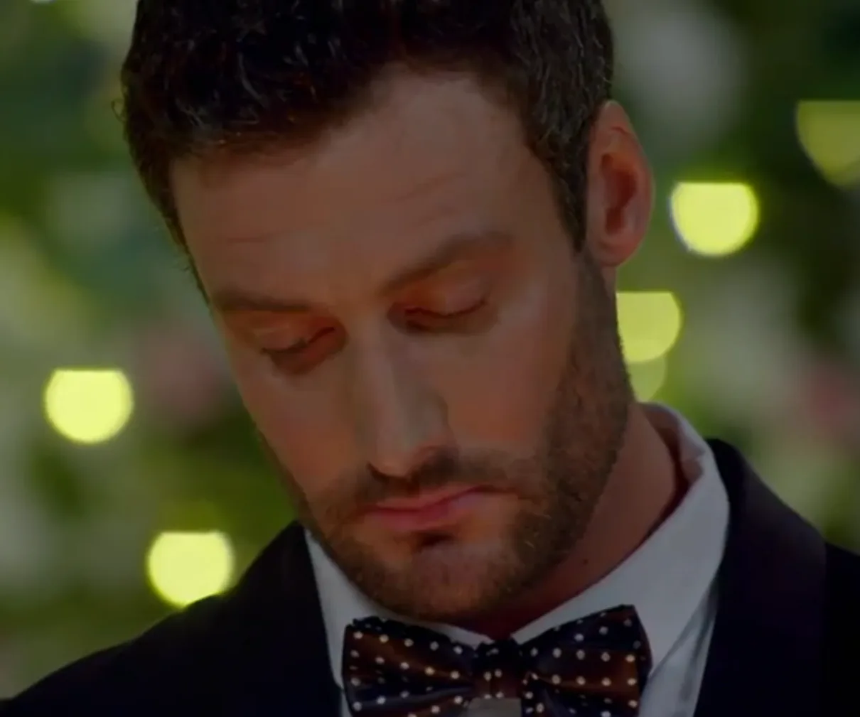 A man in a suit with a polka dot bow tie, looking down, set against a bokeh-lit background.
