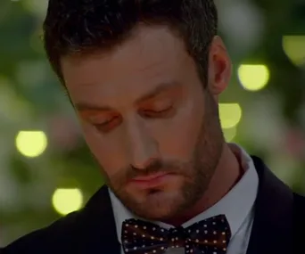 A man in a suit with a polka dot bow tie, looking down, set against a bokeh-lit background.