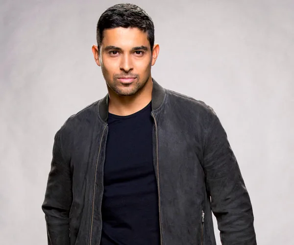 Man in a dark jacket and black shirt, posing confidently with a neutral gray background.