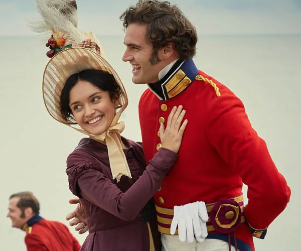 A woman in historical dress and bonnet smiles while embracing a man in a red military uniform on a beach.