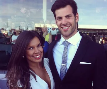 Two people posing and smiling at an outdoor event, one in a suit and the other in a white dress.