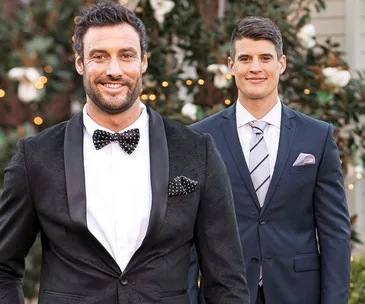 Two men in formal suits smiling, with blurred lights and foliage in the background.