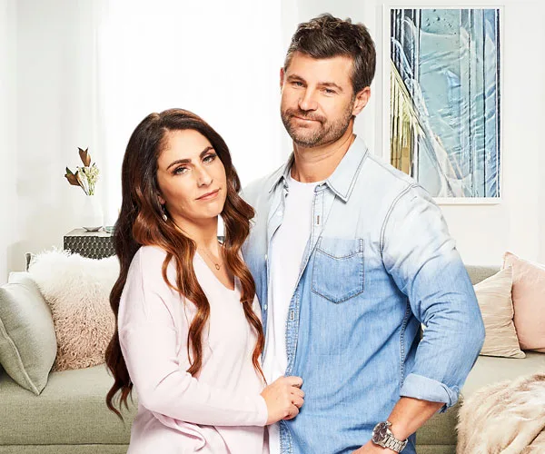 A couple standing together in a cozy, modern living room with a painting on the wall behind them.