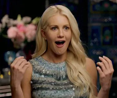 Blonde woman in blue dress appears surprised, sitting indoors with flowers in the background.