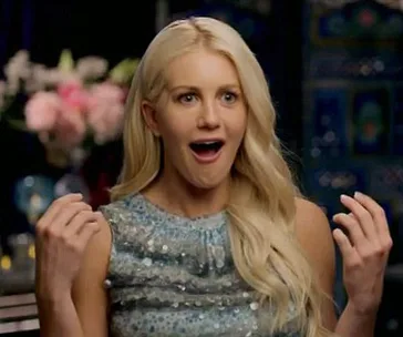 Blonde woman in blue dress appears surprised, sitting indoors with flowers in the background.