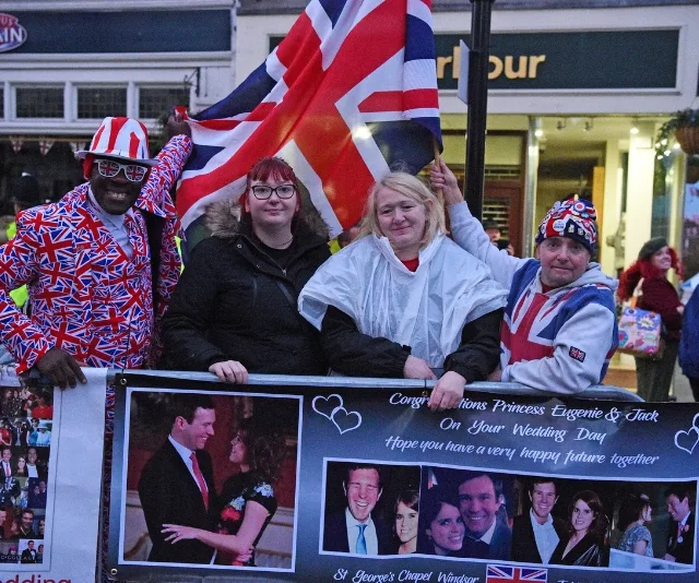 Four people with Union Jack attire holding a flag and a banner celebrating Princess Eugenie and Jack's wedding.