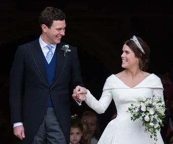 A bride and groom smile at each other while holding hands, with the bride wearing a white gown and tiara.