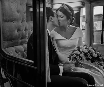 A bride and groom share a kiss inside a vintage carriage; the bride holds a bouquet and wears a tiara.