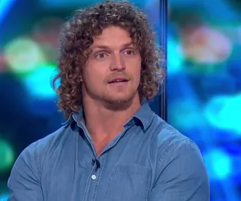 A person with curly hair in a blue shirt speaks on a talk show with a colorful background.