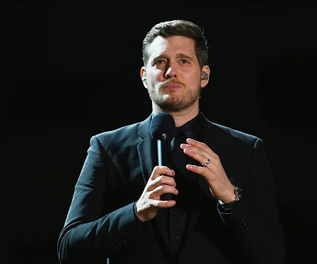 A man in a black suit holds a microphone on stage, looking thoughtful under a spotlight.