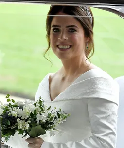 Princess Eugenie in a white wedding dress holding a bouquet, inside a car, smiling.