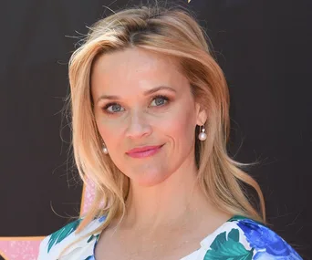 Reese Witherspoon in a floral dress, looking at the camera with a slight smile, wearing pearl earrings.