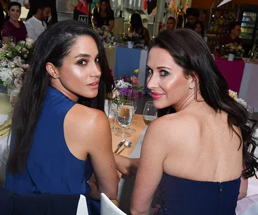 Two women in blue dresses sitting at a table, looking over their shoulders at an event with floral centerpieces.