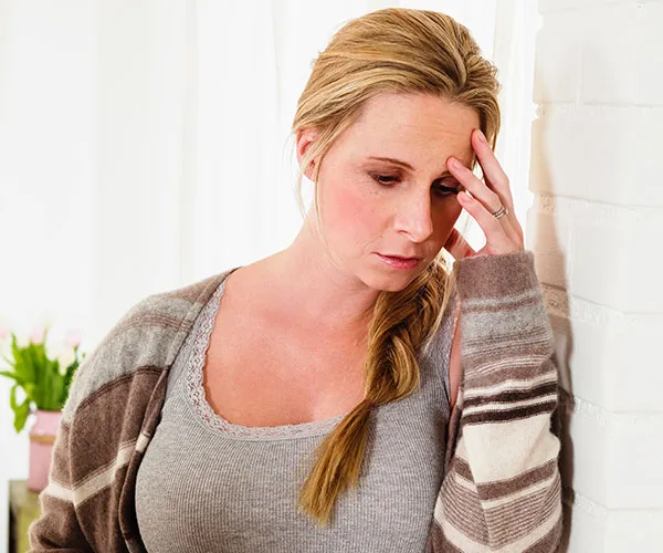 A woman with blonde hair looks down with a sad expression, leaning against a white wall, hand on forehead.