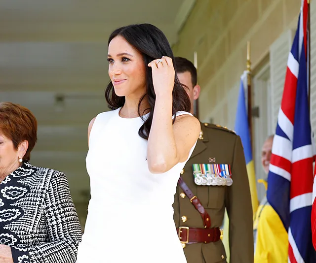 Meghan Markle in a white dress, with people and flags in the background, during a public appearance.