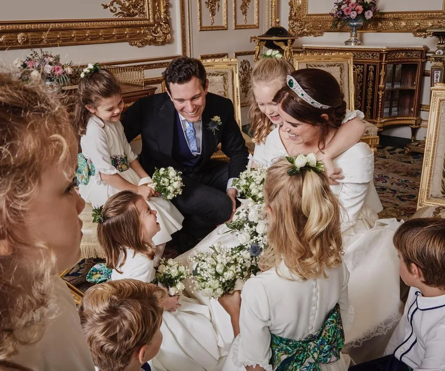 Bride, groom, and children in wedding attire gather joyfully in an ornate room with gold decor and flowers.