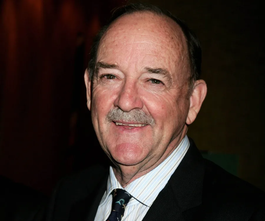 A smiling older man in a suit with a mustache, set against a dimly lit background.