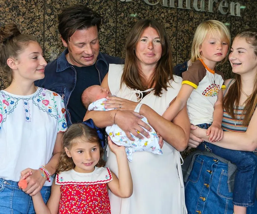 A family of six smiling together, with a mother holding a baby, standing at a public location.