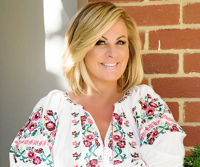 Blonde woman smiling in a floral embroidered blouse, sitting in front of a brick wall.