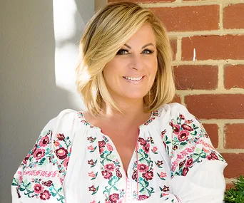Blonde woman smiling in a floral embroidered blouse, sitting in front of a brick wall.