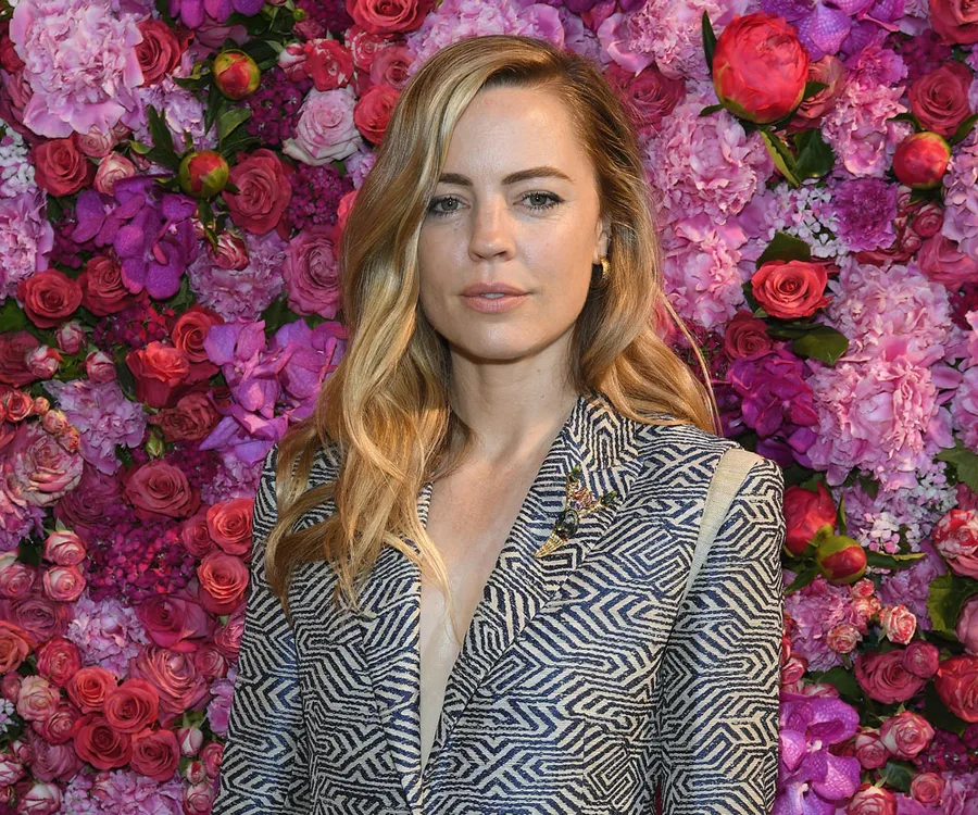 Woman in a patterned jacket stands against a vibrant pink floral backdrop.