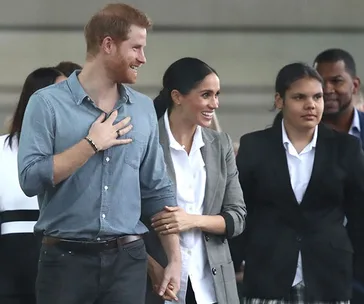 Prince Harry and Meghan Markle smiling and holding hands at a public event, surrounded by attendees.