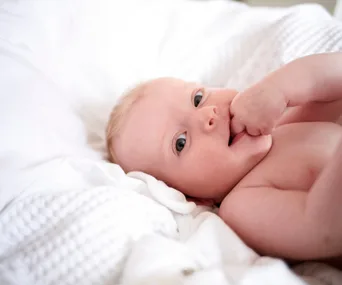 Baby lying on a white blanket, with one hand in mouth, possibly teething.