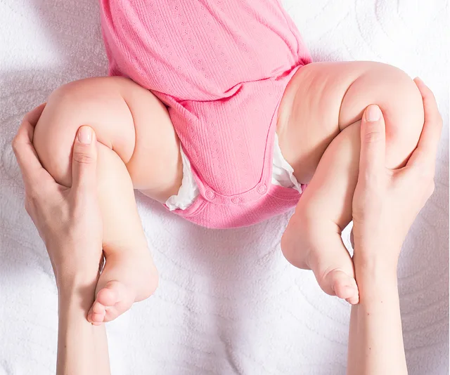 A baby in a pink outfit is held by the legs; demonstrating a hip examination technique.