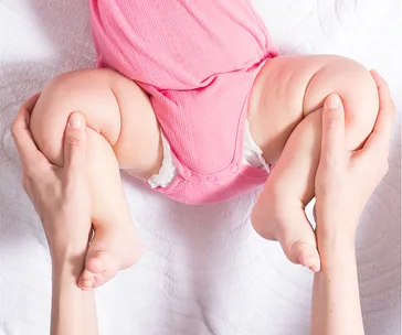 A baby in a pink outfit is held by the legs; demonstrating a hip examination technique.