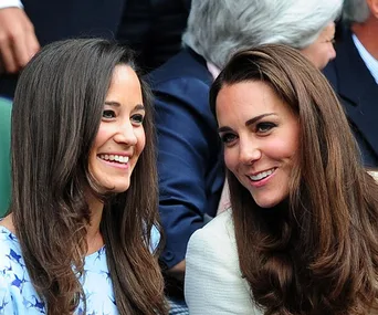 Two women with long hair smiling at each other, sitting in a crowd, likely at an outdoor event.