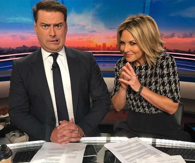 "Two news anchors on set, one with a surprised face, the other smiling mischievously; papers and coffee visible."