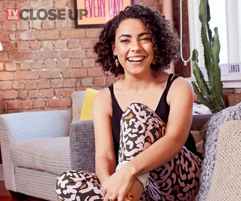 TV Week Close Up feature of a smiling woman with curly hair, wearing a black top and patterned pants, sitting on a couch.
