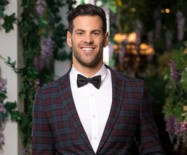Man in a plaid suit and bow tie smiling in a floral-decorated setting.