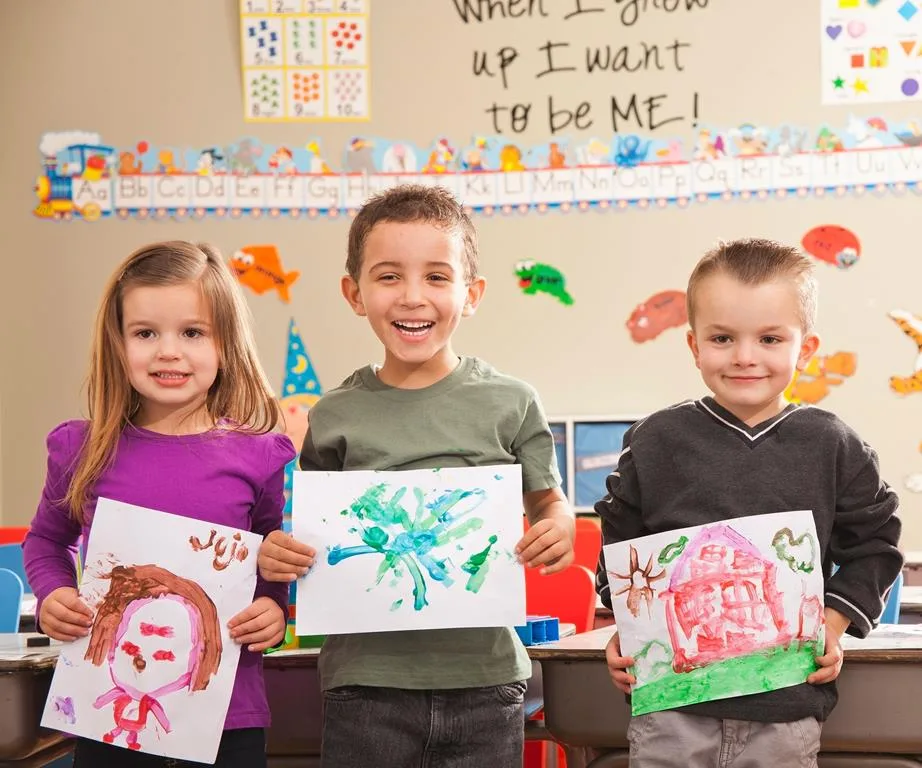 Children in a classroom proudly display their colorful paintings with smiles, under a sign that says "When I grow up I want to be ME!"