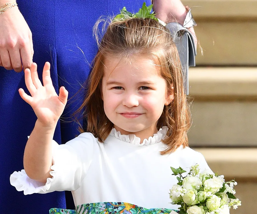 Princess Charlotte waves, dressed in white with a floral sash and holding a bouquet, standing by an adult in blue.
