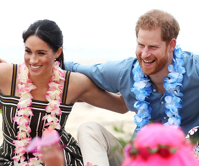 Meghan Markle and Prince Harry smiling on a beach, wearing leis, surrounded by children.