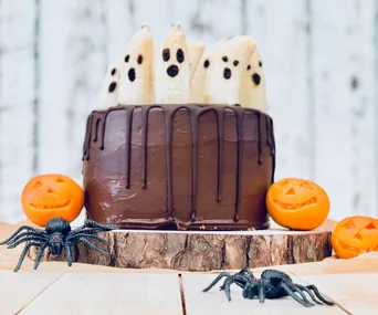 Chocolate cake with banana ghosts, surrounded by pumpkin decorations and toy spiders on a wooden base.