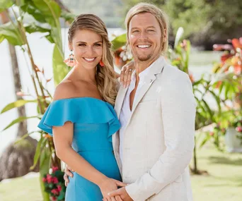 A couple smiling, the woman in a blue dress and the man in a cream suit, standing in a tropical setting.