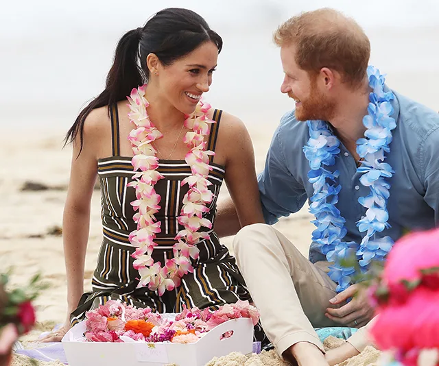 A couple wearing flower leis sits on a beach, smiling at each other, surrounded by a box of flowers.