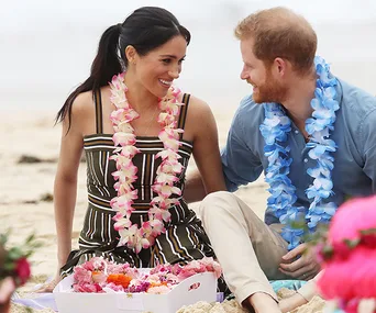 A couple wearing flower leis sits on a beach, smiling at each other, surrounded by a box of flowers.
