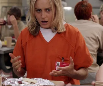 Character in orange prison uniform looks confused at a lunch table with hands gesturing over a foil-wrapped meal.