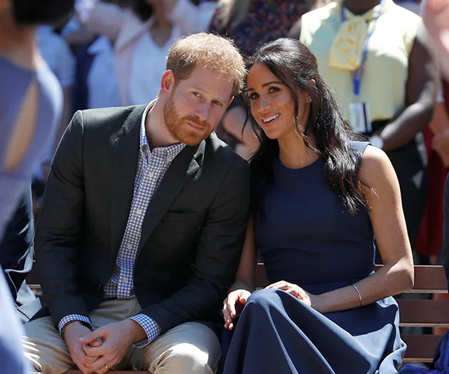 Prince Harry and Meghan Markle sitting together, smiling, at an outdoor event with a crowd in the background.