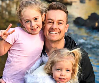 Man happily posing with two young children near a scenic water background.