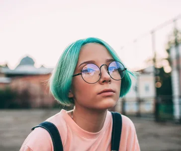 Teen with teal hair and round glasses gazes thoughtfully in outdoor setting, wearing a pink shirt and overalls.