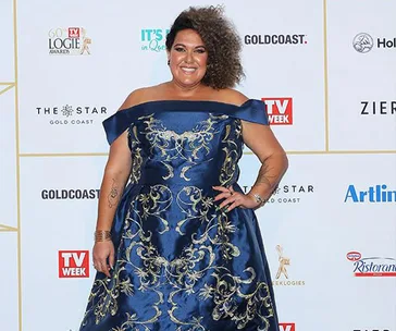 A woman in a blue off-shoulder dress with gold embroidery poses on a red carpet with TV Week and event logos in the background.