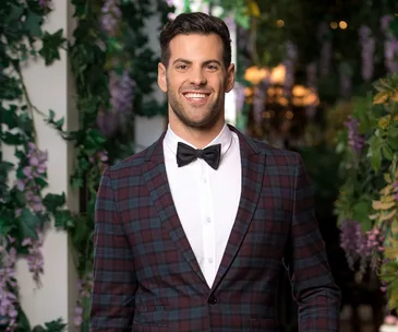 Person in a checkered suit with a bow tie smiles in a garden setting adorned with flowers and greenery.