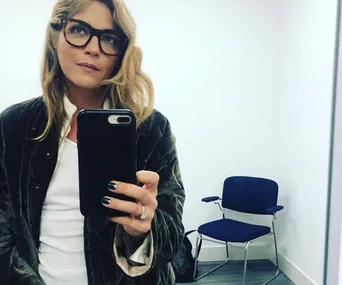 Woman with long hair and glasses takes a selfie in a room with a chair, wearing a jacket and holding a phone.
