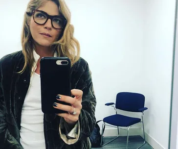 Woman with long hair and glasses takes a selfie in a room with a chair, wearing a jacket and holding a phone.
